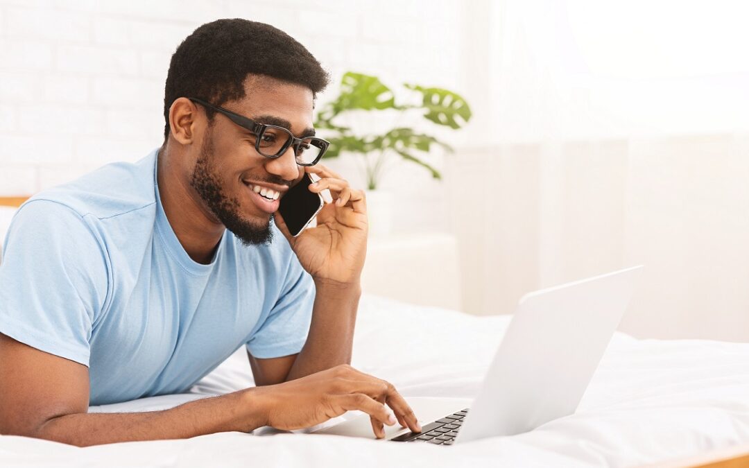 Young millennial man talking by cellphone and using laptop in bed, chatting with friends, free space