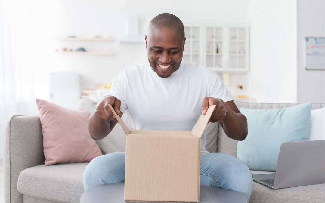 Happy black man unpacking delivery box, sitting on couch at home interior. Satisfied african american customer opening package, ordering clothes or gadgets on Internet, copy space