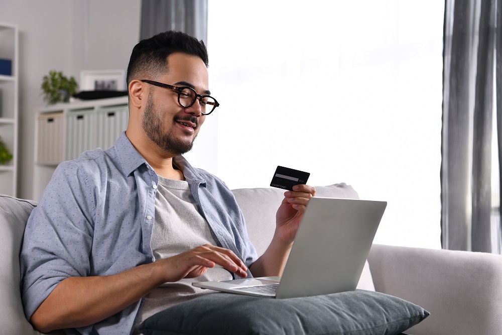 Feeling happy and confident. Young Asian man using laptop computer holding credit card for online shopping and payment at home.